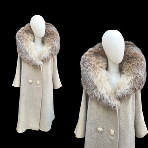 Puede incluir: Un abrigo vintage color crema con un gran y lujoso cuello de piel. El abrigo presenta un diseño cruzado con dos botones grandes y redondos. Las mangas son anchas y acampanadas. El abrigo se exhibe en un maniquí sobre un fondo negro.