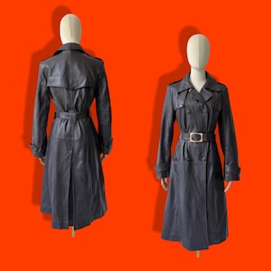 May include: A dark charcoal gray leather trench coat displayed from the front and back. The coat features a double-breasted button closure, a wide collar, shoulder epaulets, long sleeves with buckled cuffs, and a matching belt with a rectangular buckle.