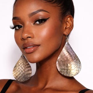 May include: Large, teardrop-shaped earrings with a textured, iridescent surface. The earrings have a silver hook and a scale-like pattern in shades of gold, silver, and purple. The earrings are worn by a person with dark skin and black hair.