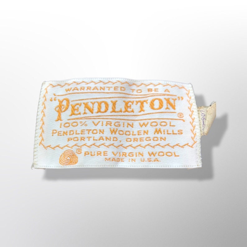 May include: A white rectangular label with the word "PENDLETON" in gold lettering. The label states "100% Virgin Wool" and "Made in U.S.A." in gold text. The label has a decorative gold border and a small tag.