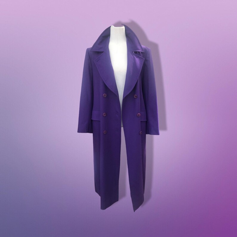 May include: A long, double-breasted purple coat with a wide collar and button closures. The coat is displayed on a white mannequin against a gradient purple background. The coat's length extends to the ankles.