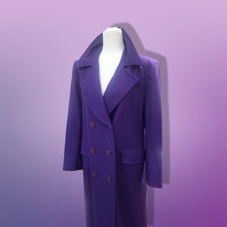 May include: A long, double-breasted purple coat with a wide collar and two front pockets. The coat has six buttons and is displayed on a white mannequin. The coat is made of a soft, textured fabric.