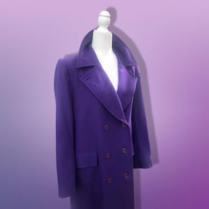 May include: A vibrant purple double-breasted coat displayed on a white mannequin. The coat features a wide collar, two rows of dark buttons, and a tailored fit. The coat is made of a soft material and has a satin lining.