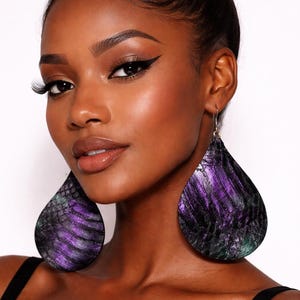 May include: Large teardrop-shaped earrings featuring a textured, snakeskin-like pattern in deep shades of purple, black, and dark green. The earrings hang from silver-toned metal hooks and have a flat, wide profile that rests against the neck.