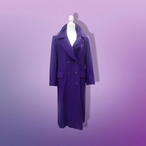 May include: A long, double-breasted purple coat with a wide collar and two rows of buttons. The coat is displayed on a white mannequin against a gradient purple background. The coat has two front pockets.