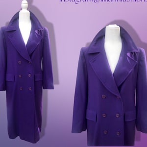 May include: A long, double-breasted purple coat displayed on a mannequin. The coat features a wide collar, two rows of buttons, and two front pockets. The coat is a solid color and appears to be made of a wool or wool-blend fabric.