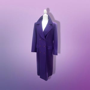 May include: A long, double-breasted purple coat with a wide collar and two rows of buttons. The coat is displayed on a mannequin against a gradient purple background. The coat has two front pockets.