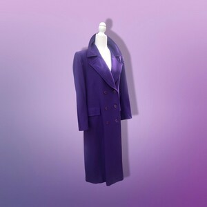 May include: A long, double-breasted purple coat with a velvet collar and lapels. The coat has a classic design with two rows of buttons and a tailored fit. The coat is displayed on a mannequin against a gradient purple background.