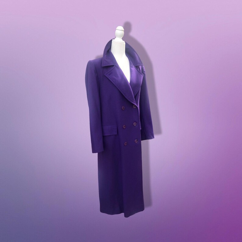 May include: A long, double-breasted purple coat with a velvet collar and lapels. The coat has a classic design with two rows of buttons and a tailored fit. The coat is displayed on a mannequin against a gradient purple background.