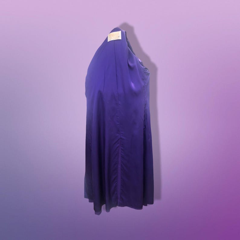 May include: A long, flowing, deep purple dress with a subtle sheen. The dress is hanging on a hanger, and a tag is visible at the top. The fabric appears to be lightweight, with a slight gathering detail down the center.