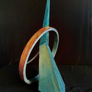 May include: A sculptural object featuring a tall, pointed teal and green triangular form intersected by a circular ring. The ring displays a textured gradient of burnt orange, copper, and gold tones, contrasting with the cool blue-green finish of the central vertical structure.
