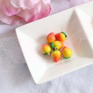 May include: Six small peach-shaped charms with green leaves and silver loops. The charms are arranged on a white plate.