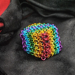 May include: A square-shaped, rainbow-colored chainmail bracelet. The bracelet is made of small, interlocking metal rings in a spectrum of colors, including red, orange, yellow, green, blue, and purple. The background is a dark, textured fabric.