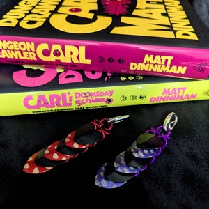 May include: Two scale maille keychains featuring overlapping metal scales in red, black, and purple, attached to silver-toned metal clips. The keychains are positioned in front of two books titled Dungeon Crawler Carl and Carl's Doomsday Scenario.