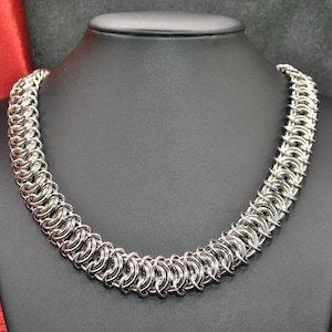 May include: A silver chain necklace made of interlocking metal rings. The necklace is displayed on a black mannequin bust.