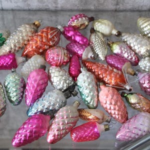 Può includere: Una collezione di decorazioni natalizie vintage a forma di pigna. Gli ornamenti sono in vari colori, tra cui rosa, arancione, argento e verde. Hanno una superficie lucida e sono disposti su una superficie riflettente.