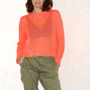 May include: A bright orange, open-weave sweater with long sleeves. The sweater is paired with olive green pants. The top has a round neckline and a loose fit. The sweater's texture is visible, with large holes creating an airy design.
