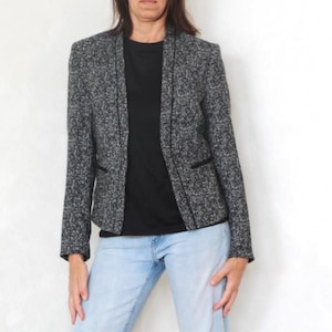 May include: A gray and black tweed blazer with a black trim and a black shirt underneath. The blazer has a classic, open-front design. The person is wearing light blue jeans.