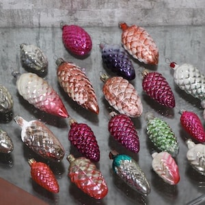Può includere: Una collezione di decorazioni natalizie vintage a forma di pigne. Gli ornamenti sono in vari colori, tra cui rosa, rosso, argento, verde e viola. Sono fatti di vetro e hanno una superficie lucida e riflettente.