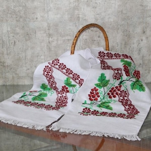 May include: A white rectangular linen runner featuring intricate cross-stitch embroidery of red berries and green leaves, finished with fringed edges. The runner is draped over a curved wooden handle, showcasing a detailed geometric border pattern.