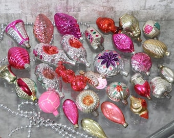 May include: A collection of vintage glass Christmas ornaments in various shapes and sizes, including spheres, bells, and teardrops. The ornaments feature metallic, glittery, and painted finishes in shades of pink, red, silver, gold, and orange.