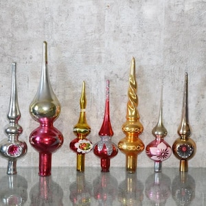 Può includere: Una collezione di sette puntali per albero di Natale in vetro vintage di varie forme e colori, tra cui argento, oro, rosso e rosa. Questi ornamenti presentano finiture riflettenti, punte affusolate e dettagli decorativi dipinti o glitterati.