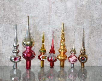 Puede incluir: Una colección de siete puntas de árbol de Navidad de vidrio vintage en varias formas y colores, incluyendo plata, oro, rojo y rosa. Estos adornos presentan acabados reflectantes, remates puntiagudos y detalles decorativos pintados o con purpurina.