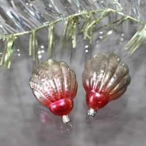 May include: Two vintage seashell-shaped Christmas ornaments. The ornaments are clear with a silver sheen and a red base. A silver tinsel garland is in the background. Each ornament has a small metal loop for hanging.