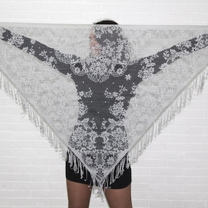 May include: A white lace shawl with a floral pattern and fringe trim. The shawl is draped over a person's shoulders.