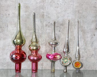 Puede incluir: Cinco puntas de árbol de Navidad de vidrio vintage dispuestas en fila. Las dos de la izquierda tienen bases esféricas rojas con agujas doradas. Las tres de la derecha son plateadas, con la del medio con detalles rosas y las otras mostrando detalles pintados coloridos similares a flores.