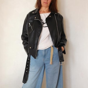 Può includere: Giacca di pelle nera con cerniere argentate e cintura, abbinata a pantaloni di jeans blu chiaro. La giacca ha un classico stile biker con colletto a revers e chiusura asimmetrica con zip. Un top bianco completa l'outfit.