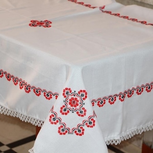 Può includere: Tovaglia bianca con ricami floreali rossi e neri. I bordi presentano un delicato bordo in pizzo. La tovaglia è drappeggiata su un tavolo, mostrando il ricamo dettagliato.
