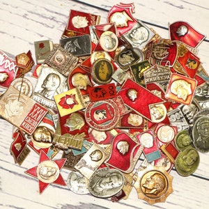 May include: A collection of vintage Soviet era enamel pins featuring red, white and gold colours. The pins feature various images including portraits, flags and symbols. The pins are in good condition and would be a great addition to any collection.