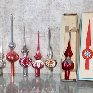 May include: A selection of vintage Christmas tree toppers in red and silver. The toppers have different shapes and decorative details. One is in its original box, which has a red and blue design.