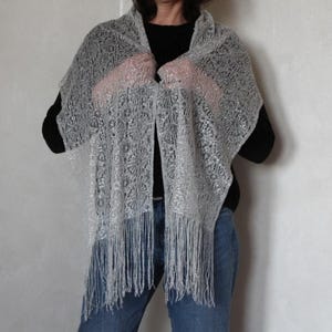 May include: A silver-grey, open-weave shawl with a delicate, lacy pattern and fringe. The shawl is draped over the shoulders, creating a soft, flowing silhouette. The intricate design and light colour suggest a formal or elegant accessory.