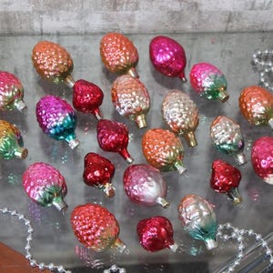 May include: A collection of vintage Christmas ornaments shaped like pinecones and berries. The ornaments are in various colors, including red, pink, orange, and green, with silver metal caps. A string of silver beads is also present.