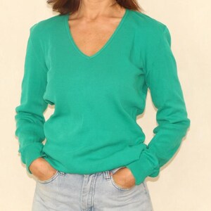 Può includere: Un maglione verde smeraldo a maniche lunghe con scollo a V. Il maglione è abbinato a jeans di denim blu chiaro. Le mani della persona sono nelle tasche dei jeans. Lo sfondo è di colore beige chiaro.