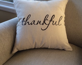 May include: A square, off-white decorative pillow with the word "thankful" printed in black cursive script. The pillow is placed on a light gray upholstered armchair, near a window.