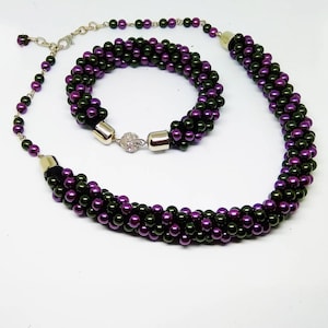 May include: A necklace and bracelet set featuring a braided design of purple and dark green beads. The necklace has a silver-toned adjustable chain. The bracelet has a silver clasp with a rhinestone accent. The jewelry is set against a white background.