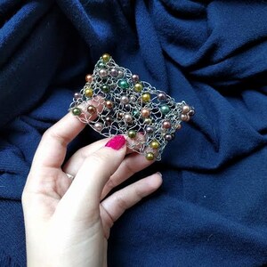May include: A silver-toned wire bracelet with a woven design, adorned with various colored beads. The beads include shades of green, gold, and brown. The bracelet is held against a dark blue fabric background.
