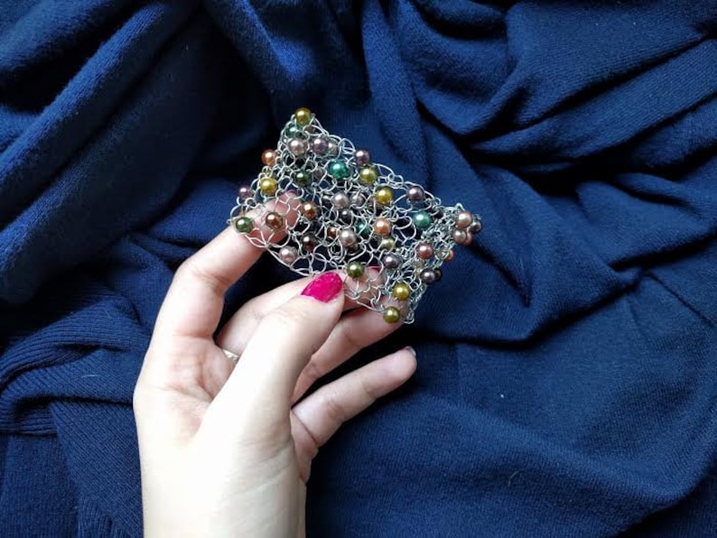 May include: A silver-toned wire bracelet with a woven design, adorned with various colored beads. The beads include shades of green, gold, and brown. The bracelet is held against a dark blue fabric background.