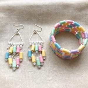 May include: A pastel-colored jewelry set featuring earrings and a bracelet. The earrings have silver triangle frames with white beads and dangling pastel cylinders. The bracelet is made of stacked pastel cylinders in pink, yellow, blue, and purple.