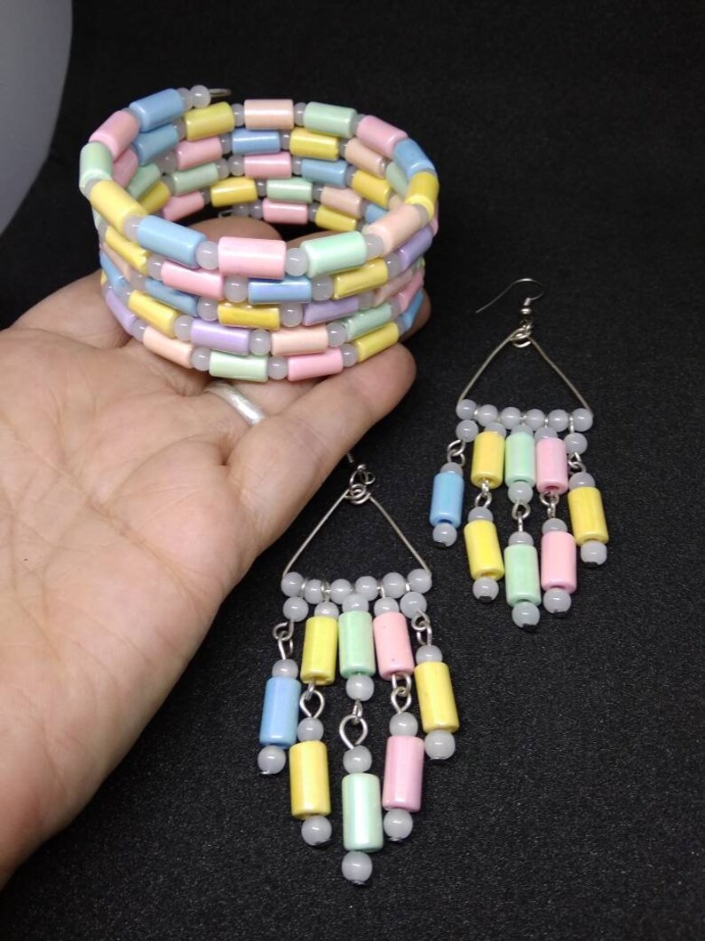 May include: A pastel-colored bracelet and earrings set. The bracelet is a coil design with rows of cylindrical beads in shades of pink, yellow, blue, and green. The earrings have a triangular wire frame with dangling beads in matching colors.