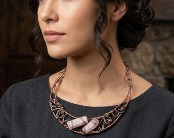 May include: A handcrafted copper wire necklace featuring two light pink quartz crystals. The necklace has an intricate, openwork design with decorative swirls and loops. The copper has a warm, reddish-brown hue, and the crystals are rectangular.