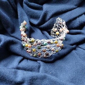 May include: A silver-toned wire bracelet with colorful beads in shades of green, yellow, purple, and brown. The bracelet is set against a dark blue fabric background, creating a contrast that highlights the jewelry's design.