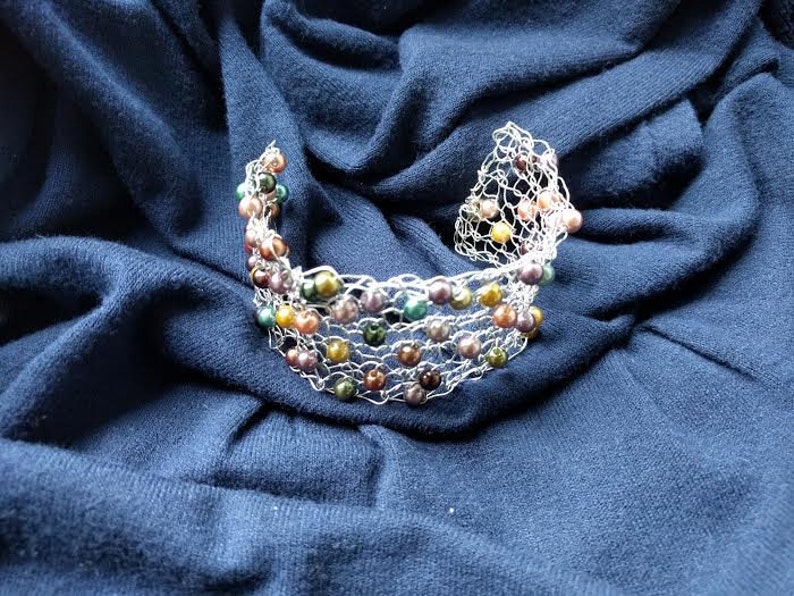 May include: A silver-toned wire bracelet with colorful beads in shades of green, yellow, purple, and brown. The bracelet is set against a dark blue fabric background, creating a contrast that highlights the jewelry's design.