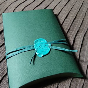 May include: A dark green, curved paper package tied with a teal ribbon and sealed with a teal wax stamp. The stamp has an embossed design. The package is on a textured, brown surface.