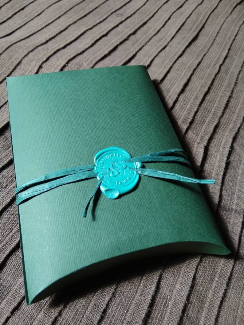 May include: A dark green, curved paper package tied with a teal ribbon and sealed with a teal wax stamp. The stamp has an embossed design. The package is on a textured, brown surface.