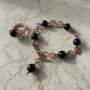 May include: A copper-toned ring and bracelet set. The ring features a single black bead. The bracelet has a series of black beads connected by copper wire with decorative swirls. The bracelet has a hook clasp and a dangling bead.