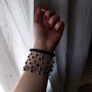 May include: A wrist adorned with two bracelets. The top bracelet is a simple black band. Below, a wider bracelet features a delicate wire mesh design, embellished with iridescent beads in shades of brown, green, and gold. The background is a sheer white curtain.
